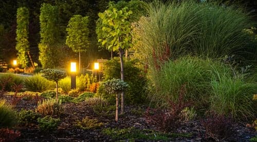 Low-voltage landscape lighting illuminating shrubs and a garden path beside a home at dusk