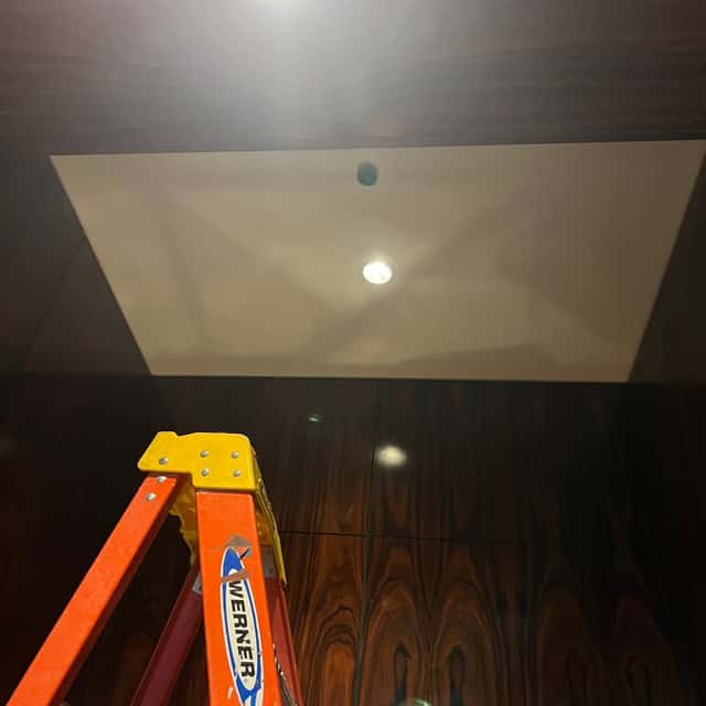Recessed downlight installed in a dark wood-panelled commercial ceiling, ladder in frame — Nebady's Electric Corporation, Palmdale, CA